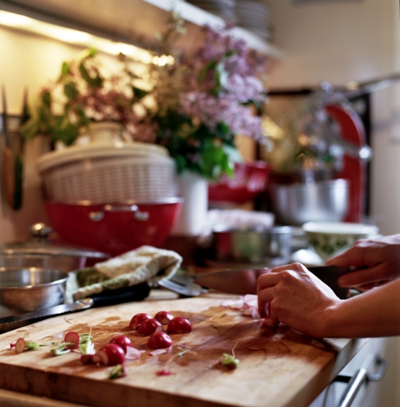 Hand chopping fresh radishes on a cutting board in a kitchen with fresh herbs and vegetables in the background.