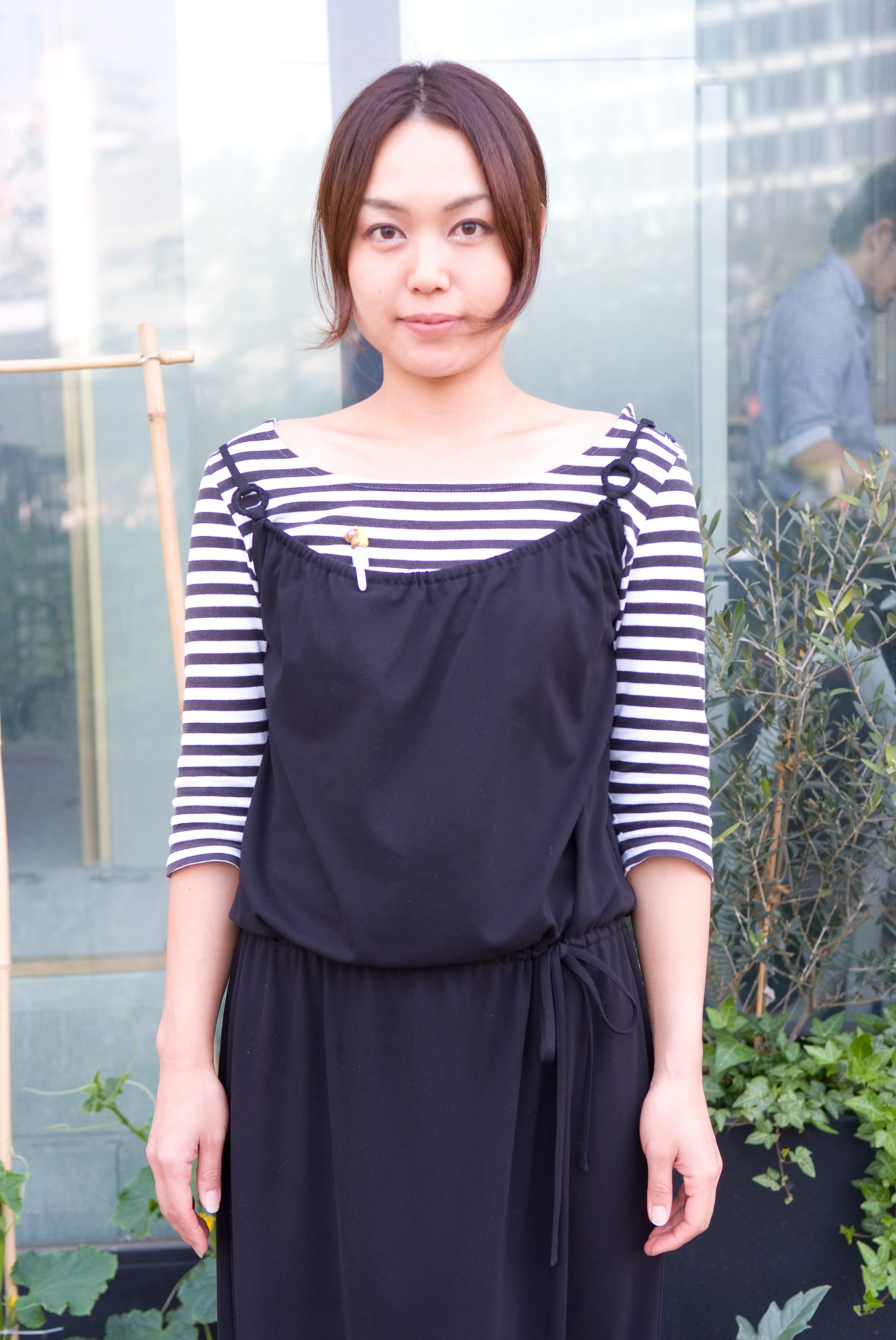 Young woman wearing a striped long-sleeve shirt and black dress standing indoors with city buildings visible in background.