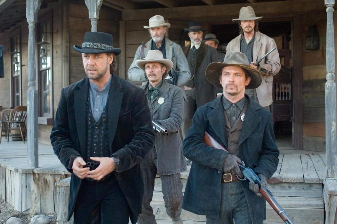 Western film scene with armed men in period clothing standing on a wooden porch.
