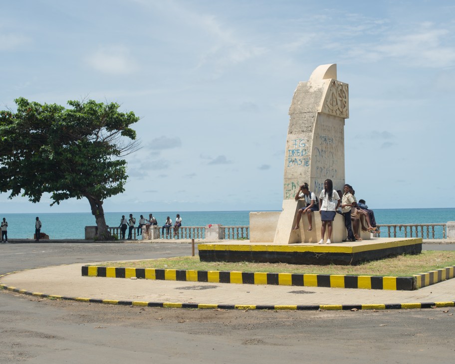 Students gathered at a waterfront monument in São Tomé with ocean and tree visible in background.