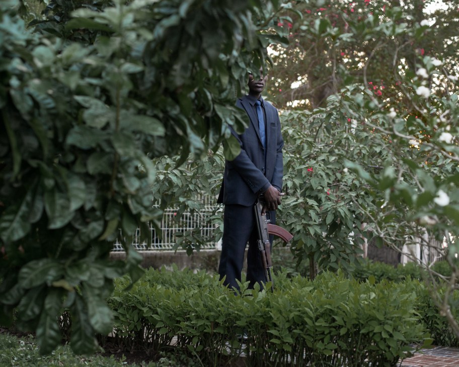 A man in a dark suit stands in a lush garden surrounded by green foliage and flowering plants.