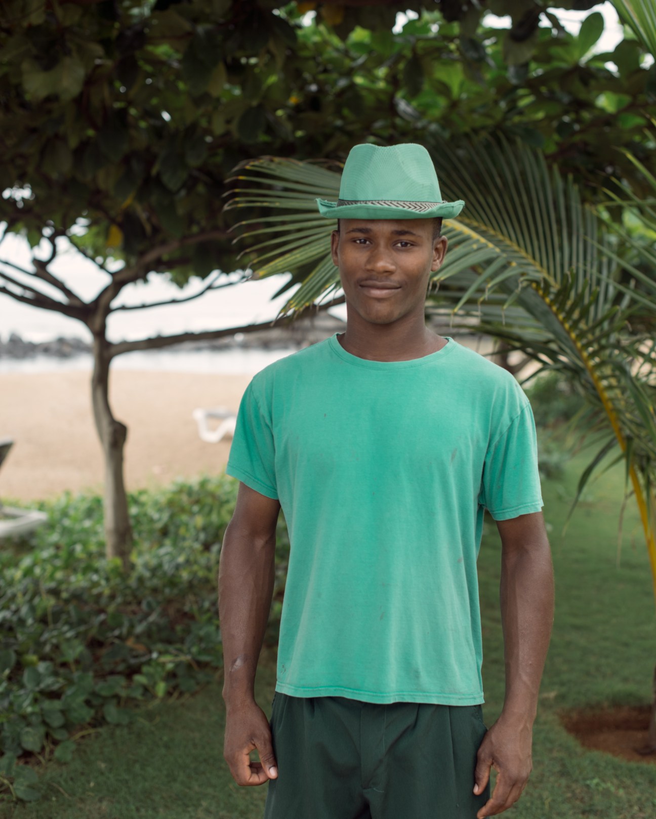 Young man wearing a turquoise hat and shirt standing among palm trees near a beach.