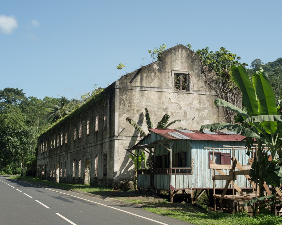 Abandoned colonial-era concrete building with barred windows alongside a deteriorated metal structure, surrounded by jungle vegetation in São Tomé.