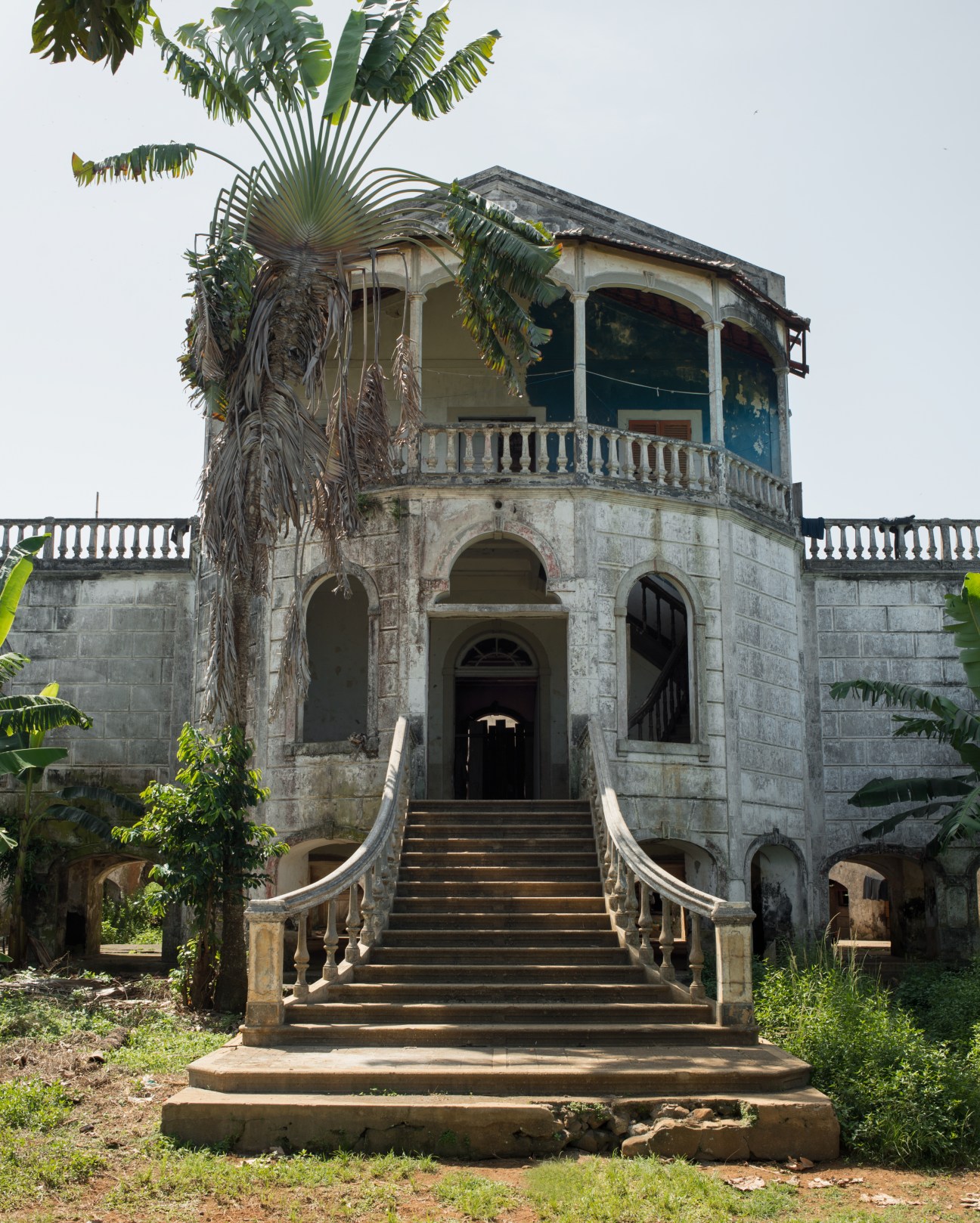 Abandoned colonial-era stone building with weathered facade, palm tree, and overgrown vegetation.