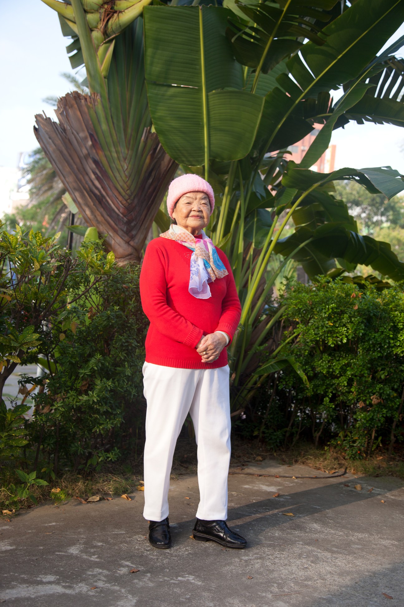 An elderly woman in a red sweater and white pants stands under large green banana leaves in a park.