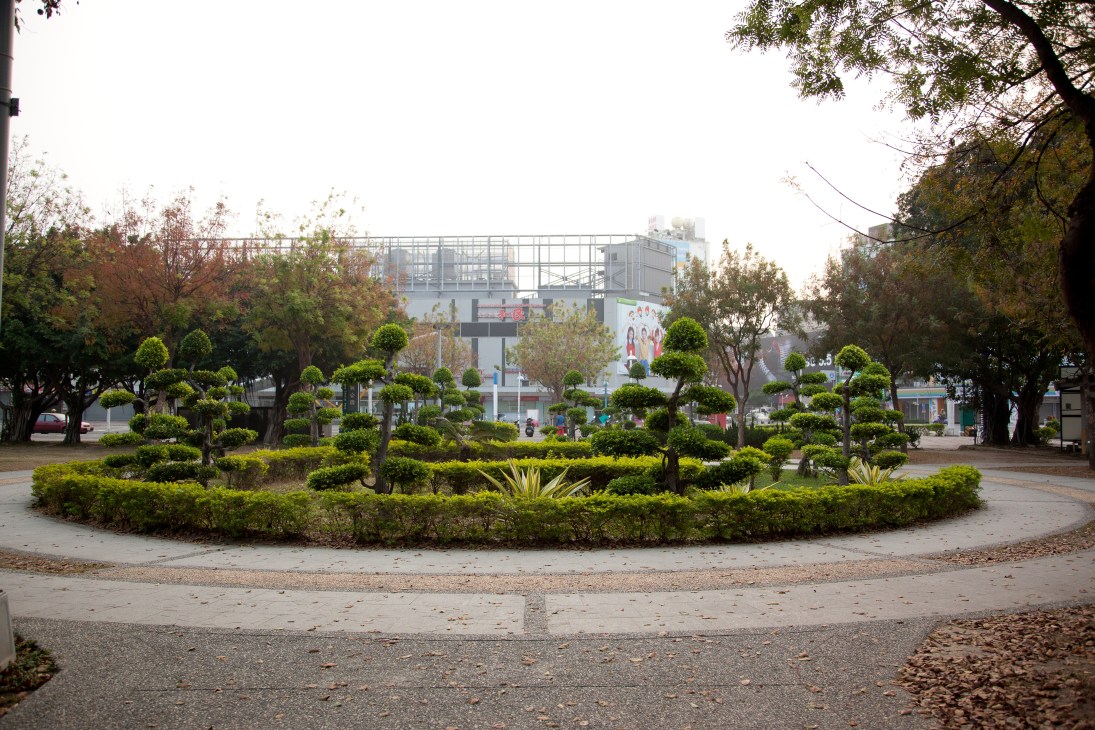 A circular roundabout in Zhongyang Park with manicured topiary shrubs, trees, and a shopping plaza visible in the background.