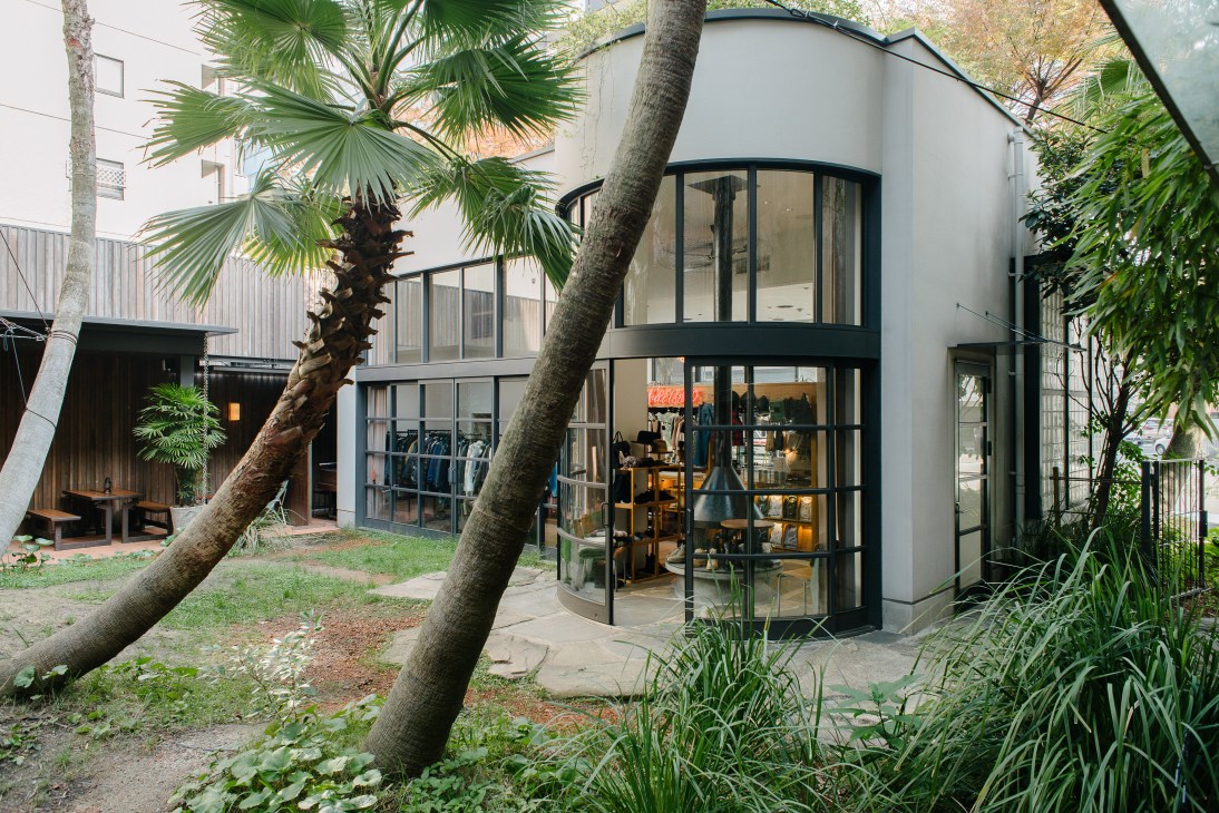 From Where I Stand kiosk at With The Style hotel in Fukuoka with modern curved glass storefront and tropical plants.