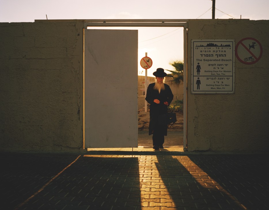 An Orthodox Jewish man with a long beard and black hat exits through a concrete gateway from a men-only beach in Tel Aviv, with signage visible on the right side.