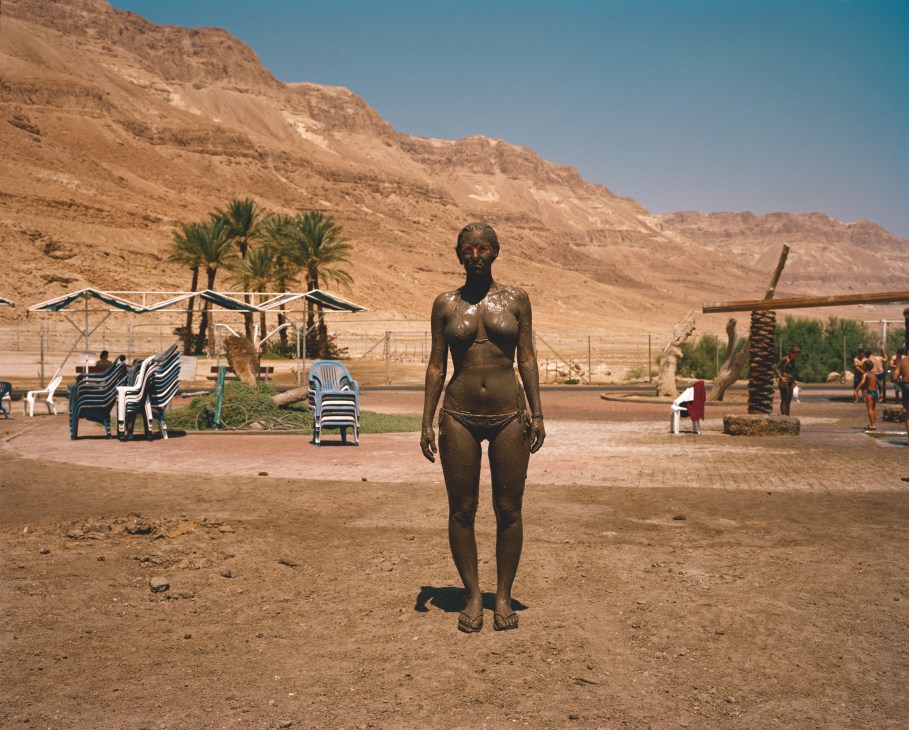 Woman covered in dark mud stands at Ein Gedi Spa near the Israel-Jordan border, with palm trees and desert cliffs behind her.