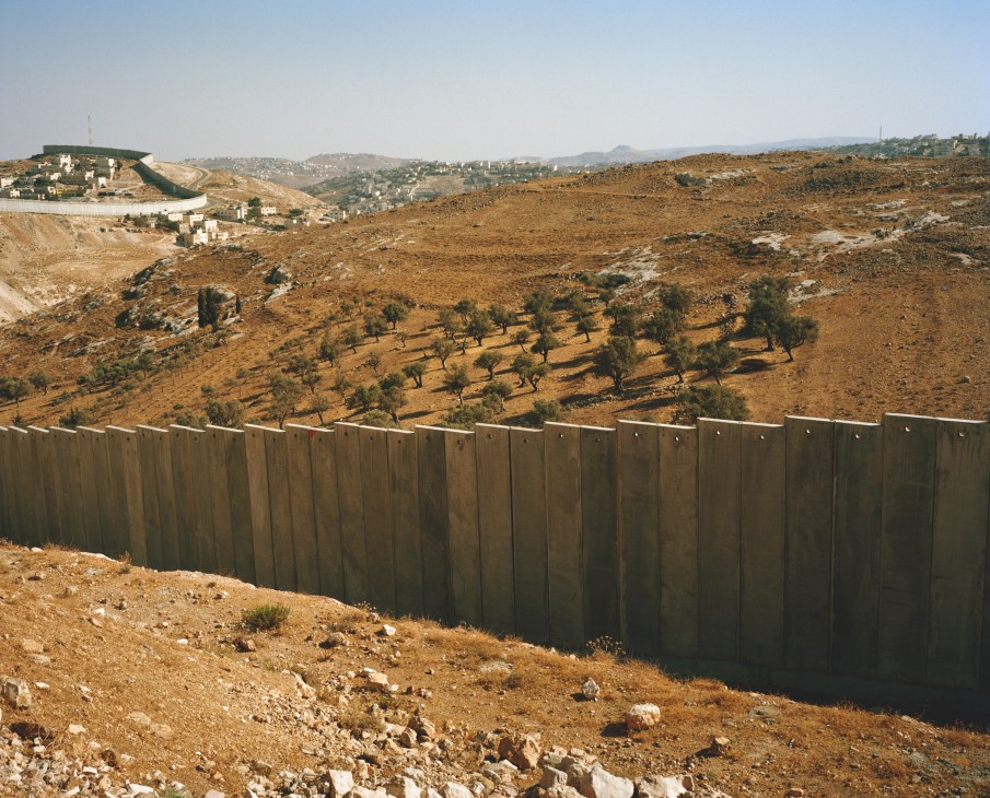 Concrete separation wall dividing West Bank landscape with olive trees and Palestinian settlements visible beyond.