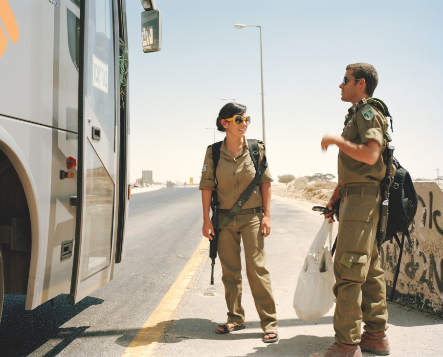 Two Israeli Defense Force soldiers in khaki uniforms stand beside a bus in the Negev desert.