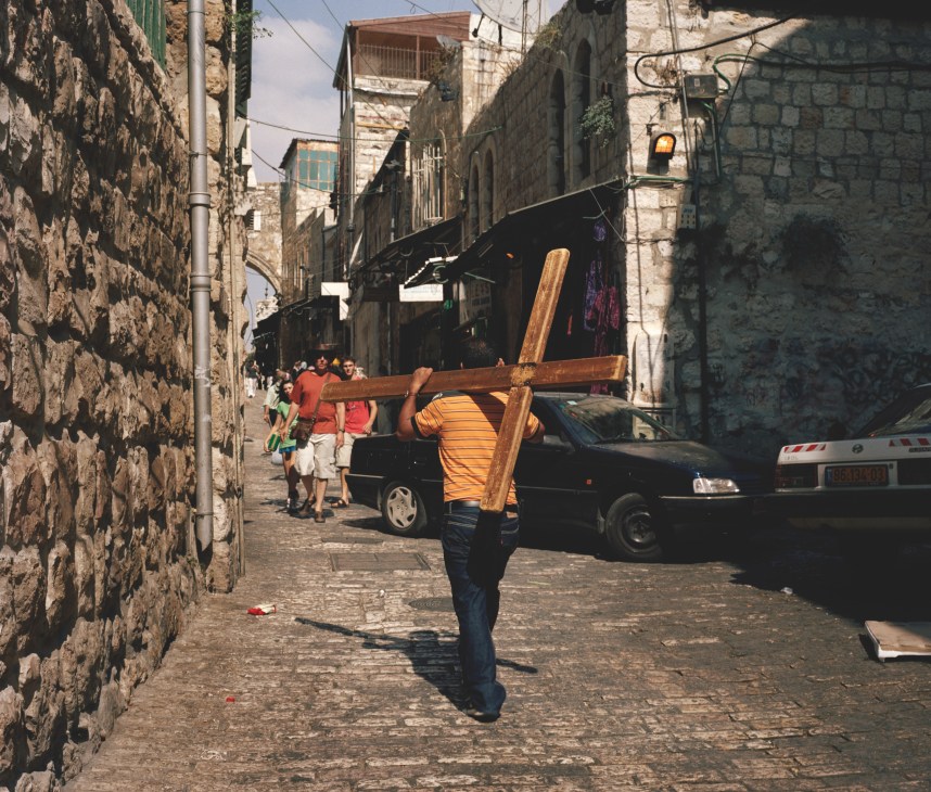 A man carries a large wooden crucifix through a narrow stone-walled street in old Jerusalem.