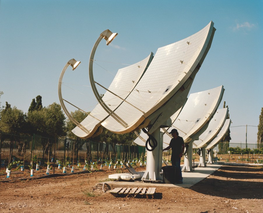 Solar panels at kibbutz Kvutzat Yavne with a person standing beneath curved reflective surfaces in a desert agricultural setting.