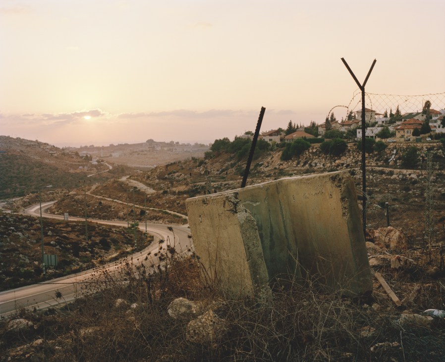 Concrete barrier with barbed wire fencing overlooking a West Bank settlement at sunset.