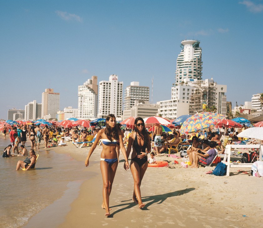 Beachgoers in swimwear walk along a crowded Tel Aviv public beach with colorful umbrellas and high-rise buildings in the background.