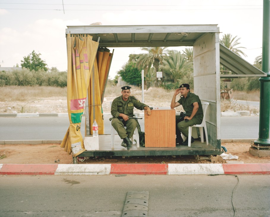 Two Palestinian soldiers in military uniforms sit in a small concrete shelter at a checkpoint entrance with yellow curtains and painted curb.
