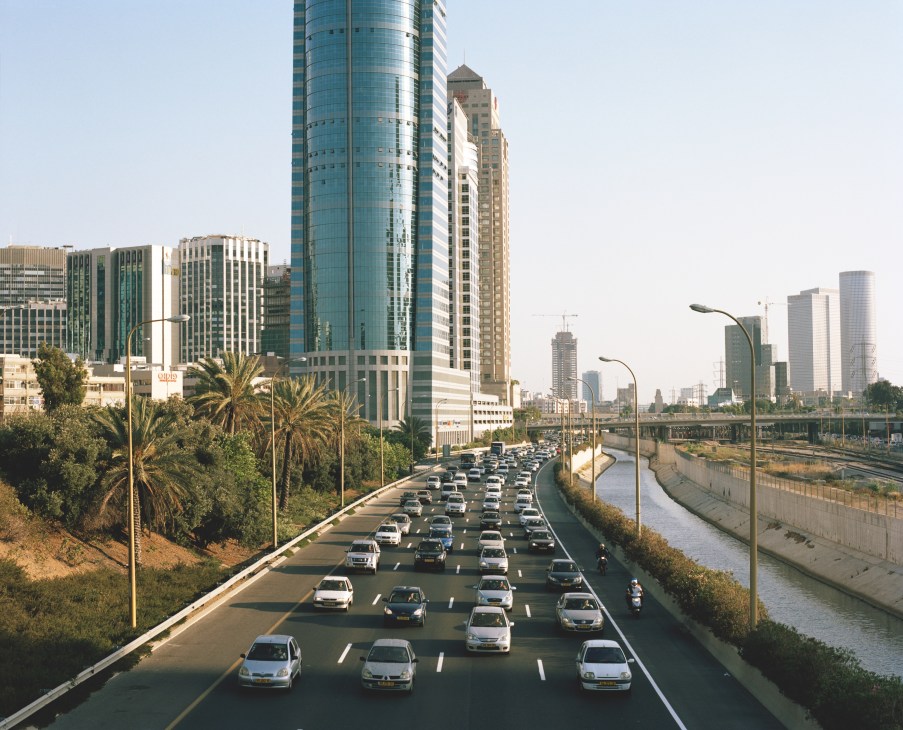 Multi-lane highway with heavy traffic leading into Tel Aviv's skyline of modern skyscrapers and palm trees.