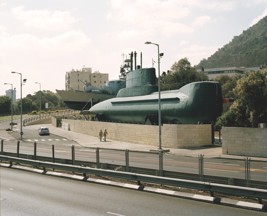 A submarine and naval vessel displayed at Haifa naval base with residential buildings and forested hills in the background.