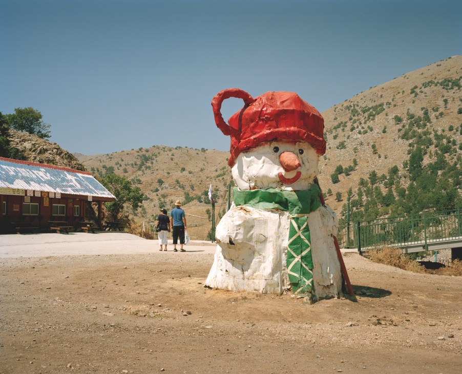 A whimsical snowman figure with red hat and green scarf stands in a desert landscape near a rustic cabin.