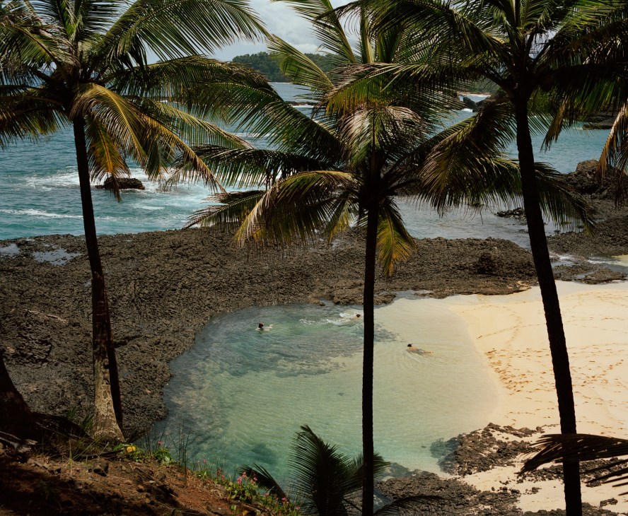 Palm trees frame a tranquil turquoise lagoon and sandy beach with calm waters on Praia Piscina, São Tomé.
