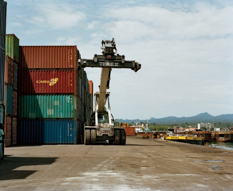Container handling crane stacking colorful shipping containers at São Tomé port with forested mountains in background.