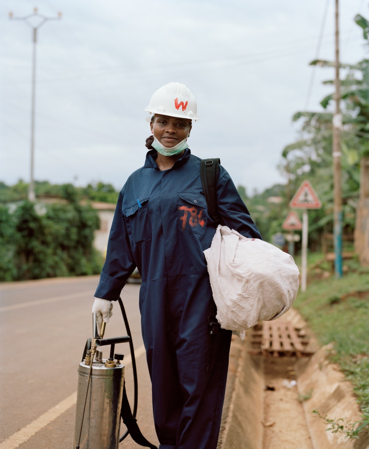 Woman in white hard hat and blue work suit holding spray equipment on a rural road.