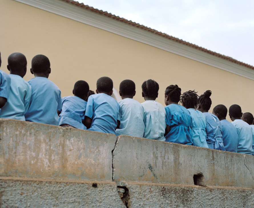 Schoolchildren in blue uniforms stand together against a concrete wall outside a church building.