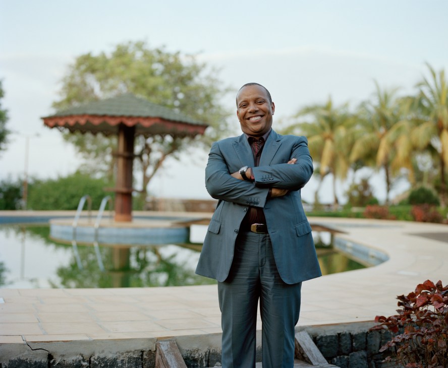 Prime minister Patrice Trovoada stands smiling with arms crossed in botanical gardens near a pool and palm trees.