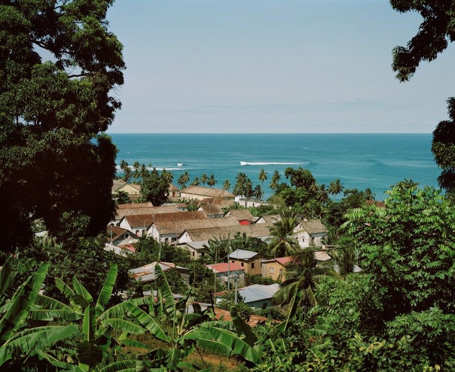 Tropical coastal village with colonial-era buildings surrounded by lush vegetation and palm trees overlooking turquoise ocean.