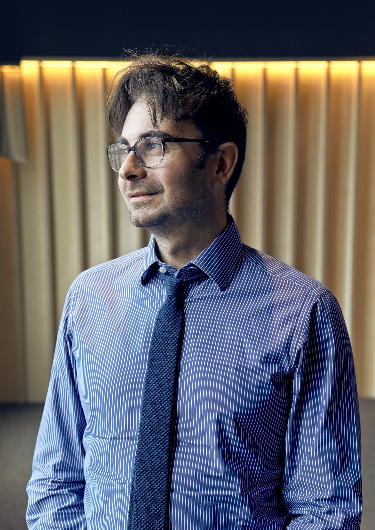 Man wearing glasses and a blue striped shirt with tie against a curtained background.