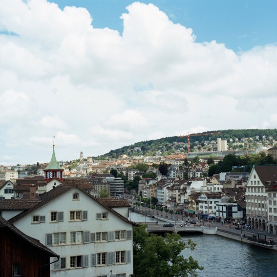 Aerial view of Zürich showing riverside buildings, church spire, and hillside residential neighborhoods under blue sky with white clouds.
