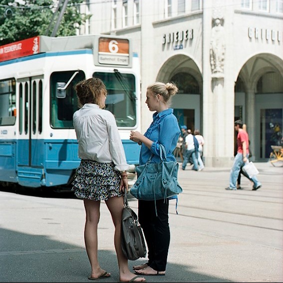 Two women stand on a Zürich street beside a blue and white tram, with historic architecture visible in the background.
