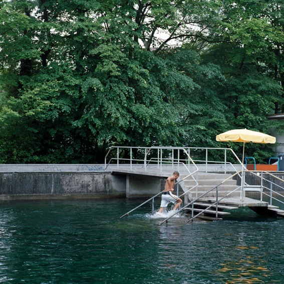 Person swimming in a river from a metal dock with yellow umbrella under lush green trees.