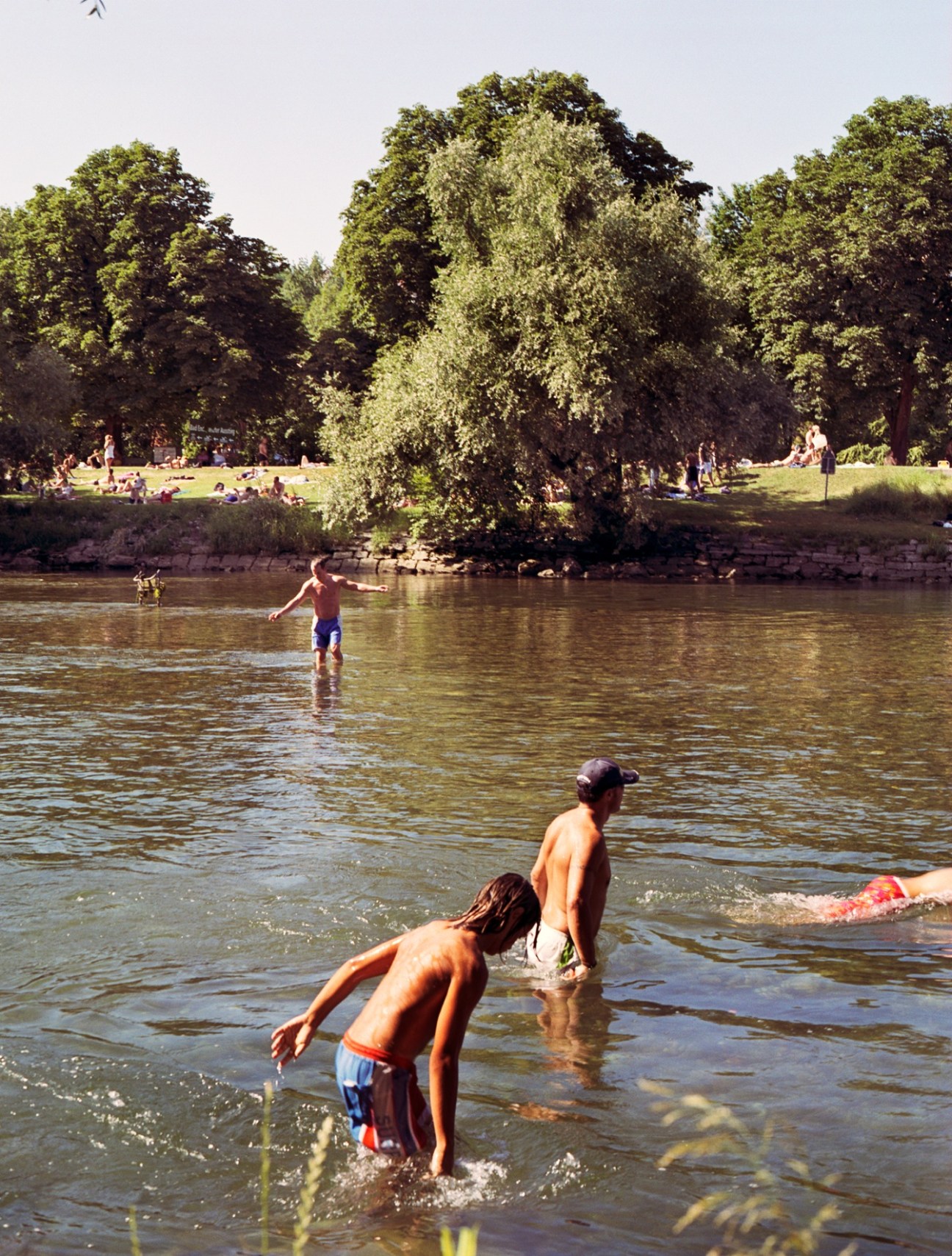 People swimming and wading in a river with tree-lined banks on a sunny day.