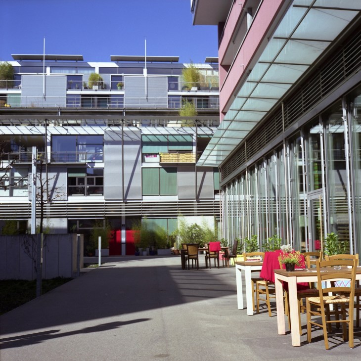 Modern courtyard with contemporary buildings, outdoor seating with red accents, and glass structures under blue sky.
