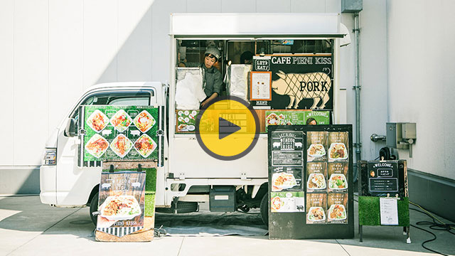 Food truck with green and white exterior displaying menu boards and packaged food items with a large yellow circle logo.