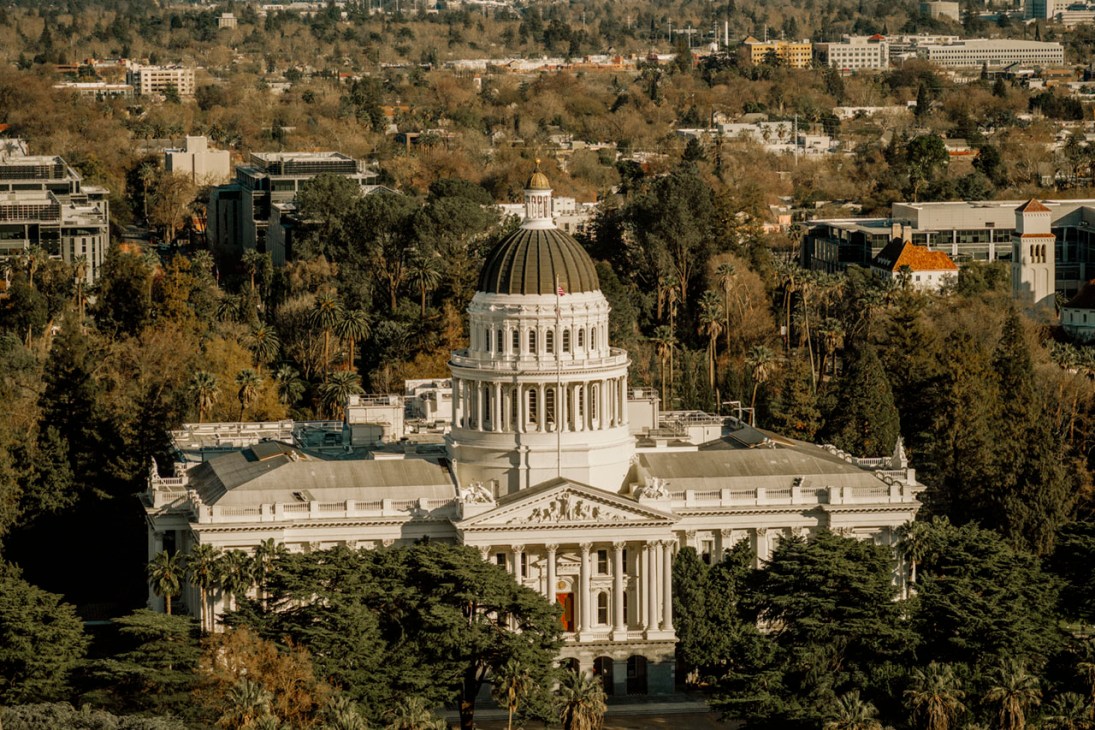 Aerial view of the California State Capitol building with its distinctive white dome surrounded by trees and urban development.