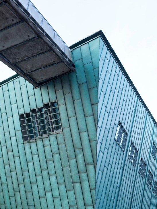Modern glass and concrete building facade with geometric window patterns and overhanging structure.