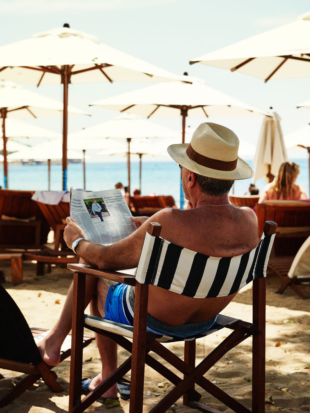 Man in striped swimwear and straw hat reads newspaper while seated on beach chair at Astir Beach.