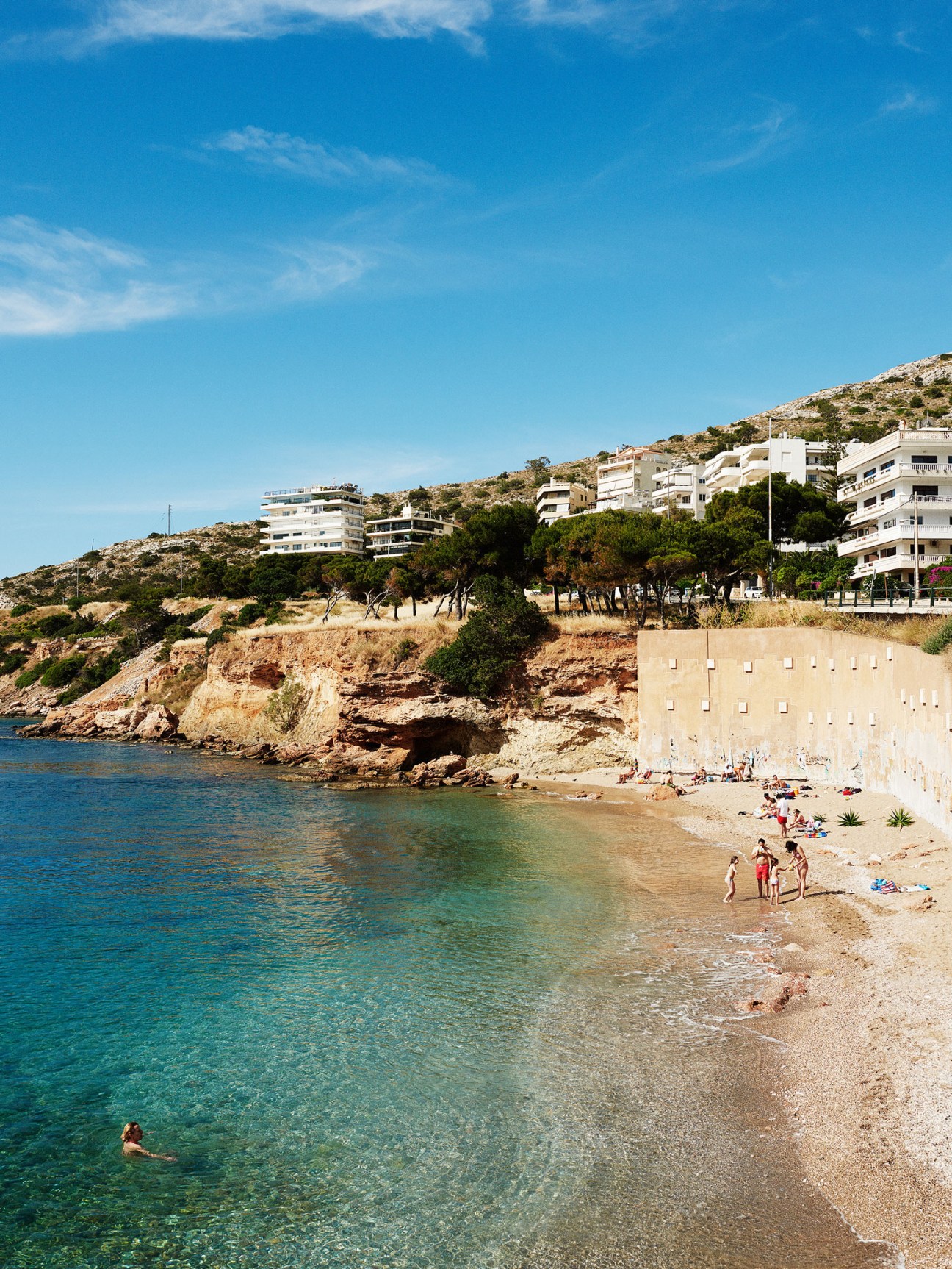 Swimmers and beachgoers enjoy clear turquoise waters at a sandy cove with white modern apartment buildings built into the rocky hillside above.